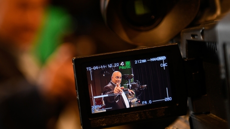 Pictured through the lens of a video camera, John Ward wears a suit and points toward the camera. 