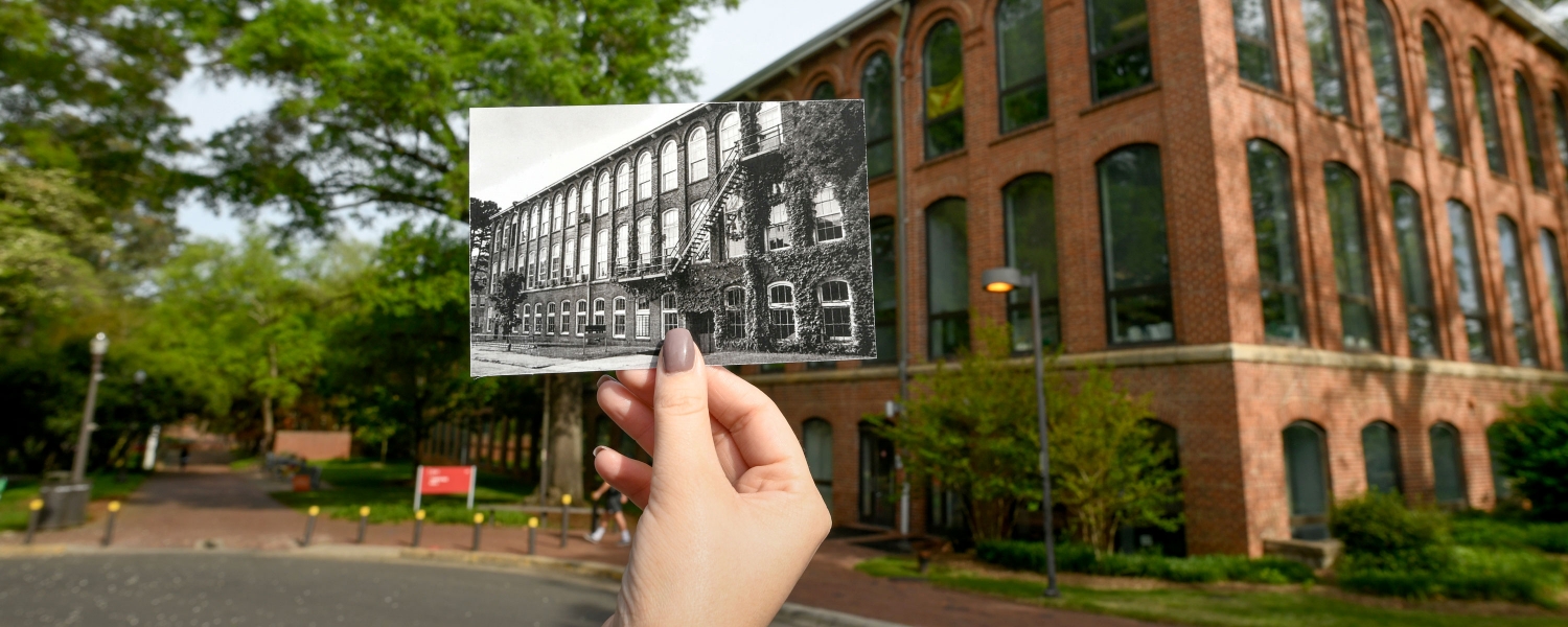 A hand holds up an archival photo of NC State's Tompkins Hall, in front of the Tompkins Hall of today.