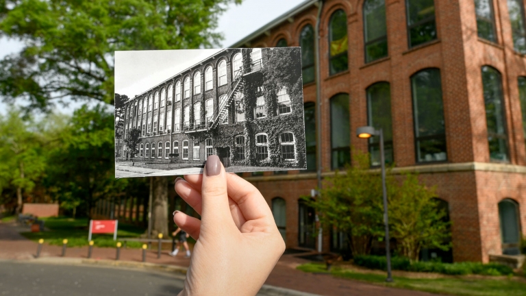 A hand holds up an archival photo of NC State's Tompkins Hall, in front of the Tompkins Hall of today.