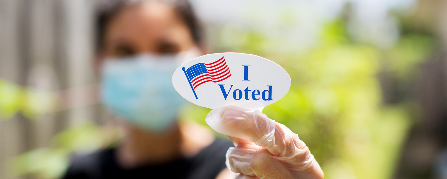 A masked person holds up a sticker that reads, "I voted."