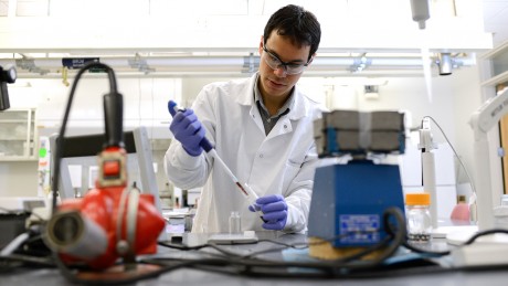 A young man in a lab coat uses a pipette in a lab setting.