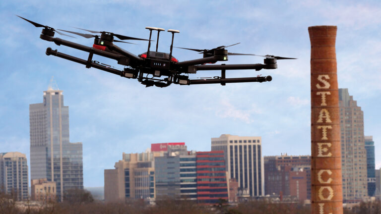 A drone flies above NC State's campus, with downtown Raleigh on the horizon.